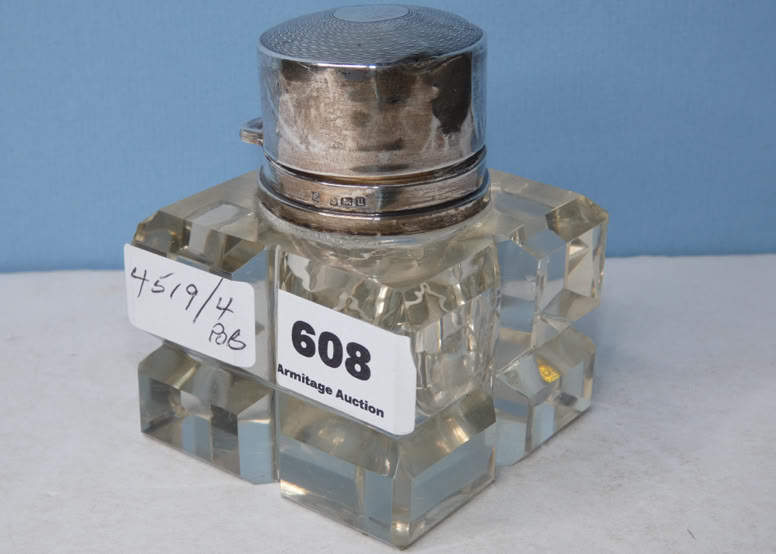 A square glass inkwell with a metal lid, featuring labels with numbers and text on its side, set against a blue background.
